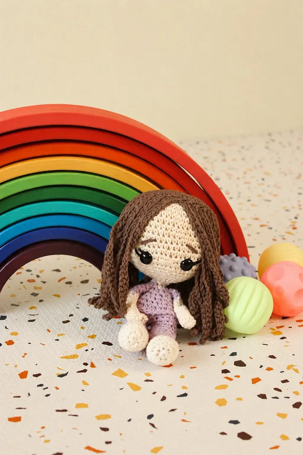 Muñeca tejida a mano sentada frente a un arcoíris de madera y pelotas sensoriales de colores