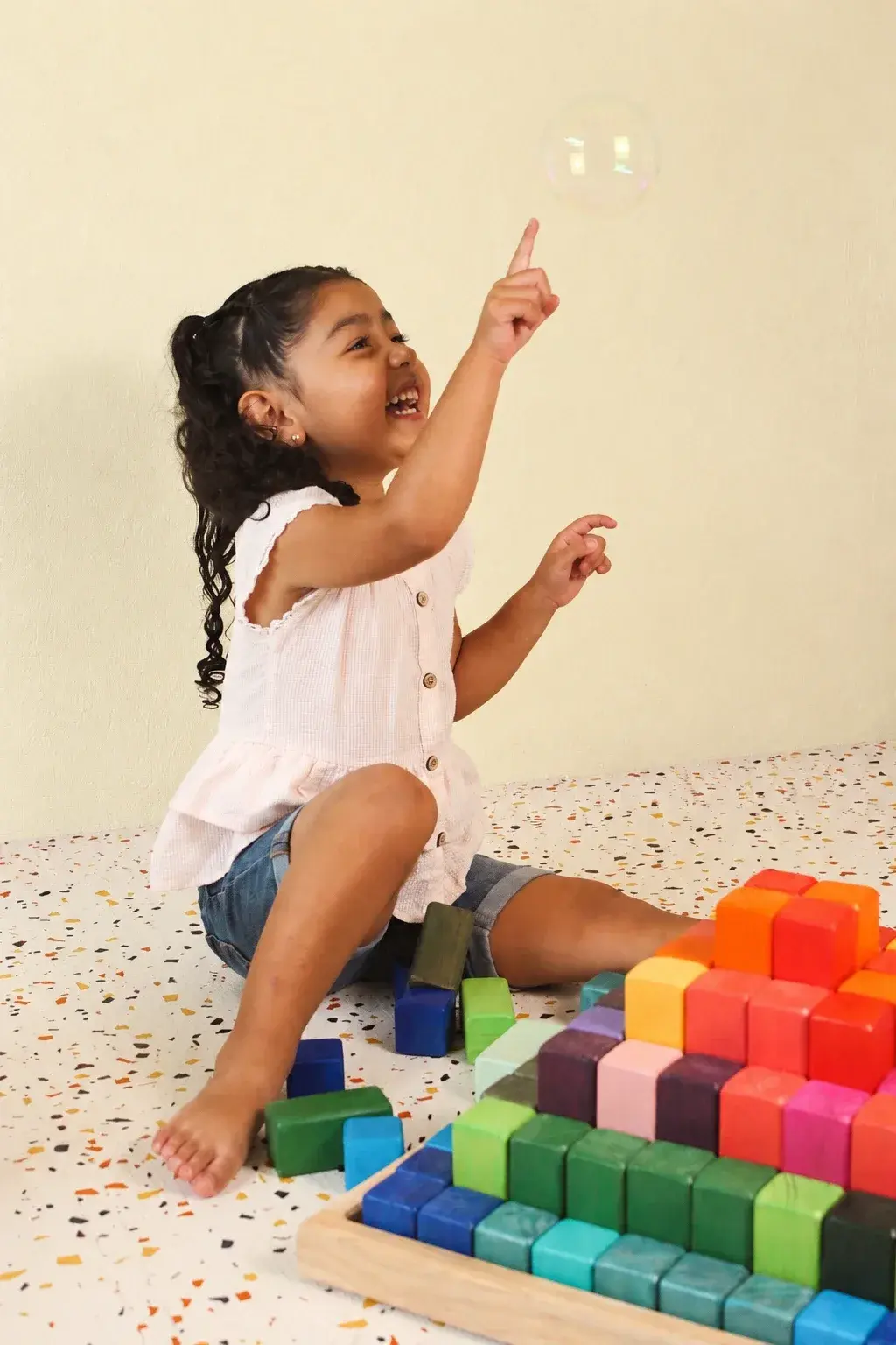 Niña riendo mientras señala una burbuja, sentada junto a una pirámide de bloques de colores